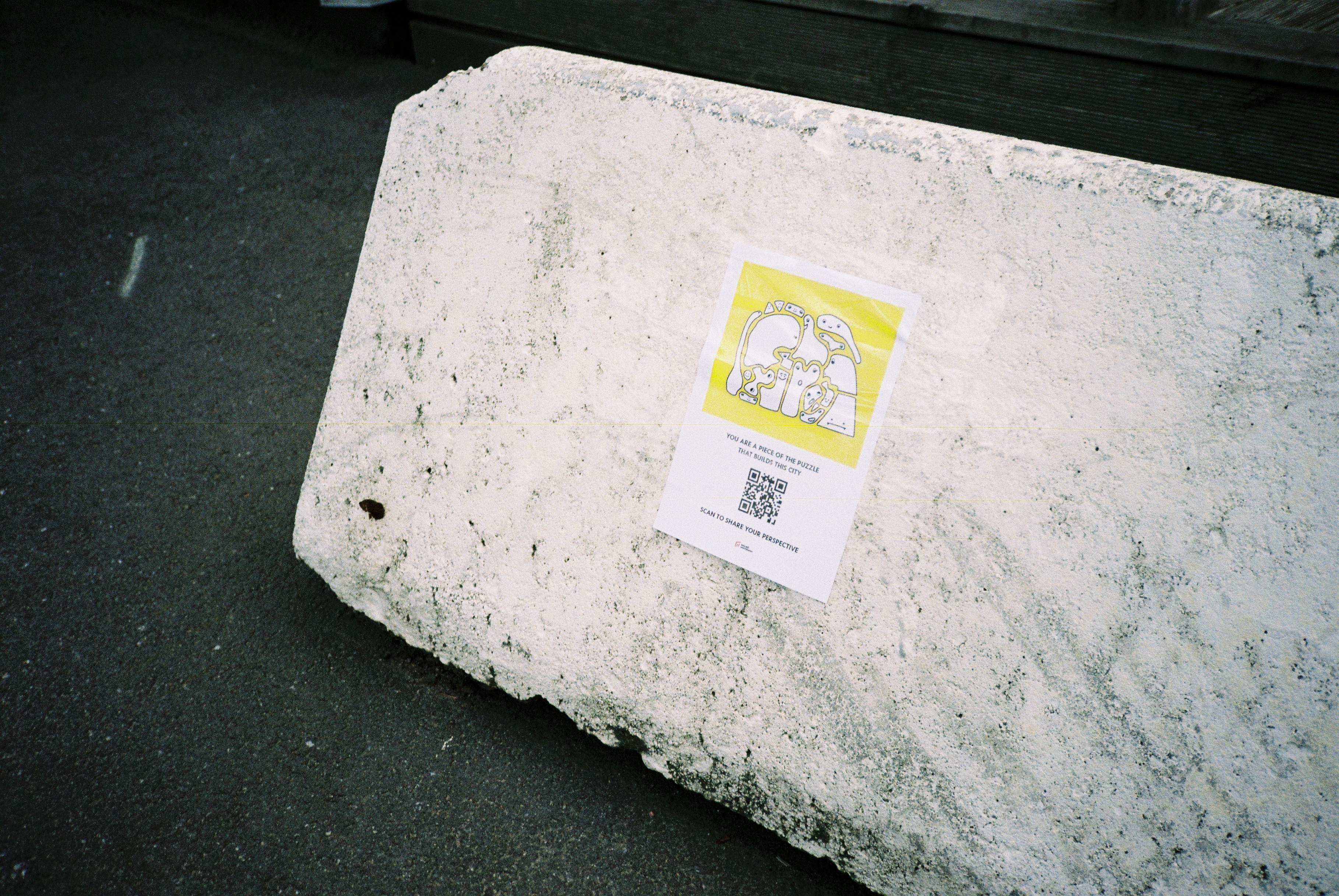 A yellow flyer with a QR code and a hand-drawn puzzle-piece illustration, taped to a concrete block at Ringön. The text reads 'You are a piece of the puzzle that shapes this city — scan to share your perspective.'